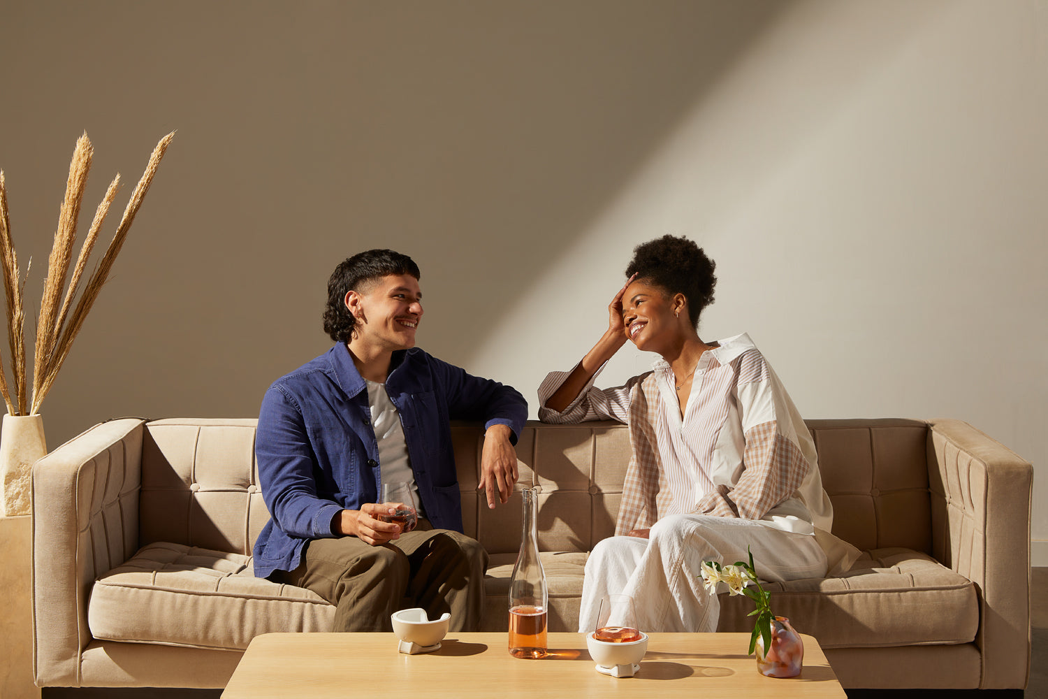 A man in a blue shirt and a woman in a white shirt sit on a tan couch. On a table in front of them sit two Stemless Wine Chillers and a bottle of rosé wine.