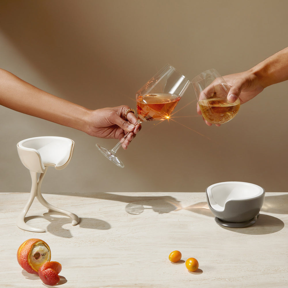 A woman holds a stemmed glass of rosé and a man holds a stemmless glass of white wine, and they clink glasses to cheers. Below, a Stemmed Wine Chiller and Stemless wine chiller sit on a neutral table with fruit and flowers.
