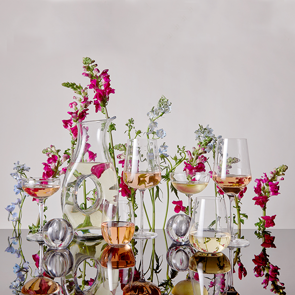 Several stemmed and stemless wine glasses and a decanter, all containing rosé and white wine, sit on a mirrored surface. They are all covered in colorful flowers.